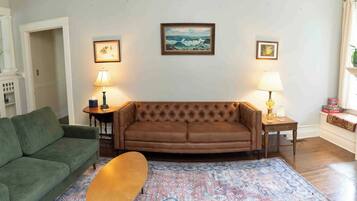 A living room with a brown sofa, a green armchair, a wooden coffee table, and a patterned rug. There are two lamps on side tables and a framed painting on the wall.