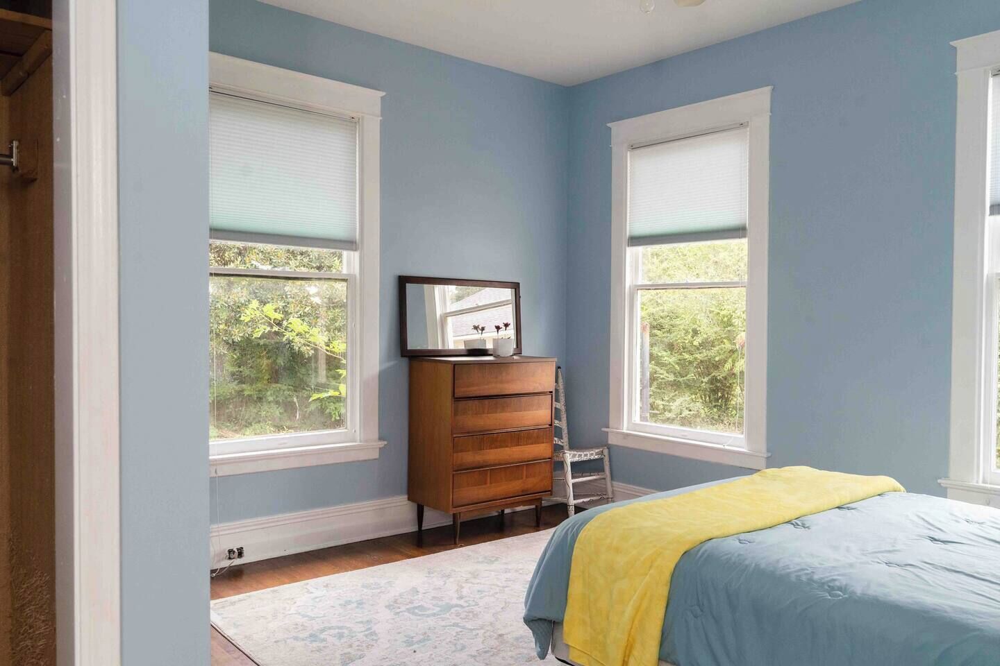 Une chambre avec deux fenêtres, une commode, un lit recouvert d’une couverture jaune et un mur blanc.