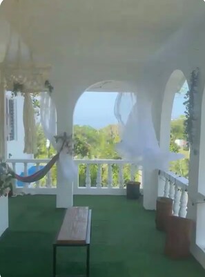 A covered outdoor area with a hammock, a bench, and a view of greenery.