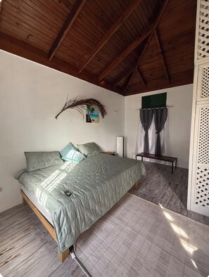 A bedroom with a wooden ceiling, a bed with green bedding, a nightstand, a bench, and a wall-mounted picture.