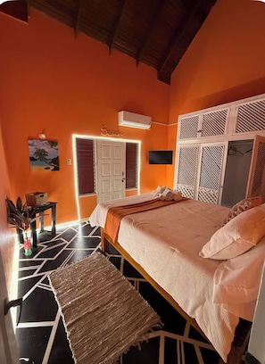 Ein Schlafzimmer mit einem Bett, einem Nachttisch, einem an der Wand befestigten Fernseher und einer Tür zu einem weiteren Raum.