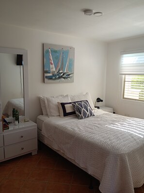 A bedroom with a bed, a dresser, a nightstand, a painting, and a window with blinds. - Delightful Zen 2-bedroom condo with ocean views in charming Añasco (Añasco)