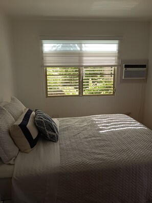 Ein Schlafzimmer mit einem Bett, Kissen, einem Fenster mit Jalousien und einer Klimaanlage.