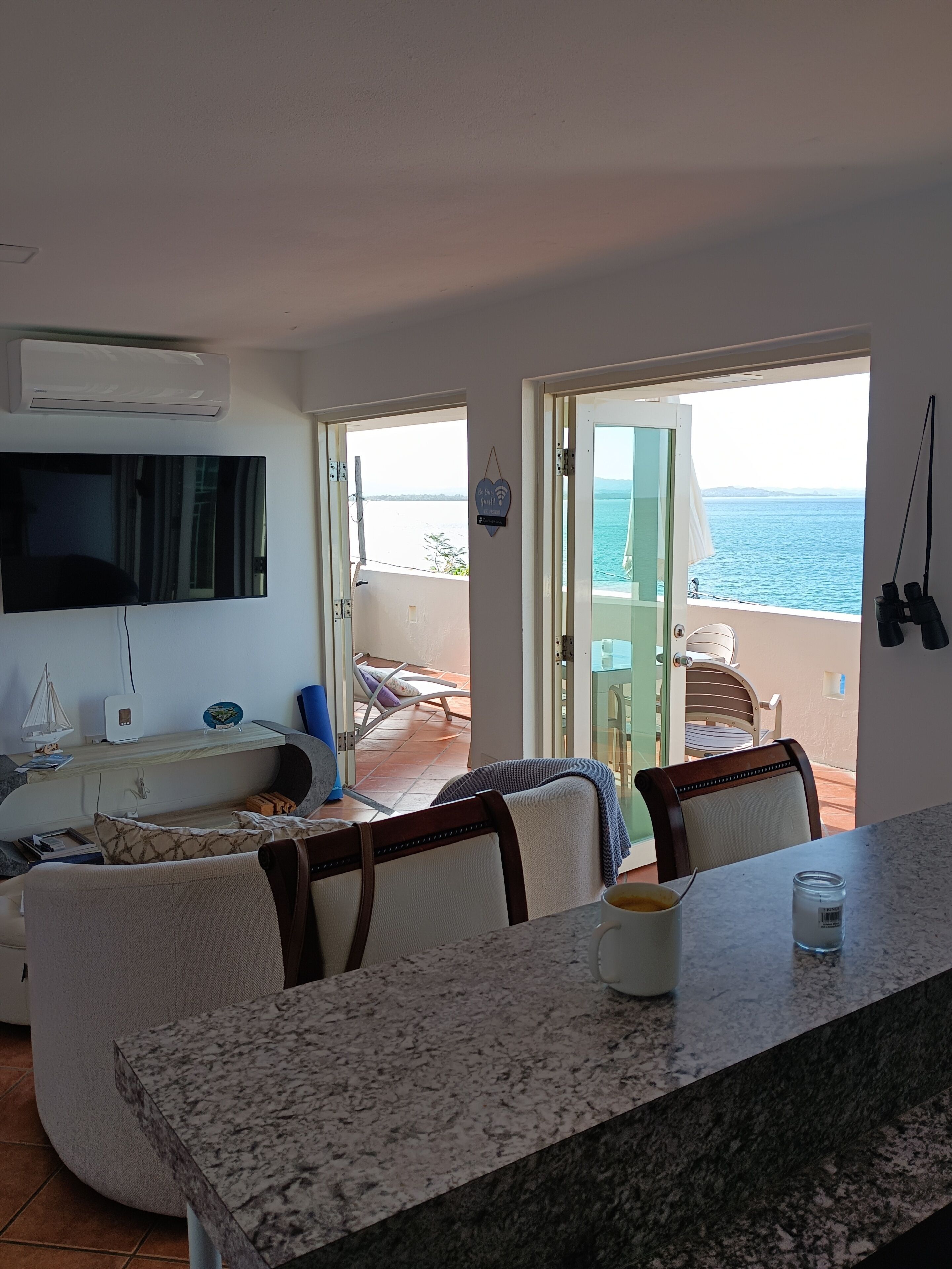 A room with a TV, a dining area with chairs, a table, and a view of the sea through an open door.