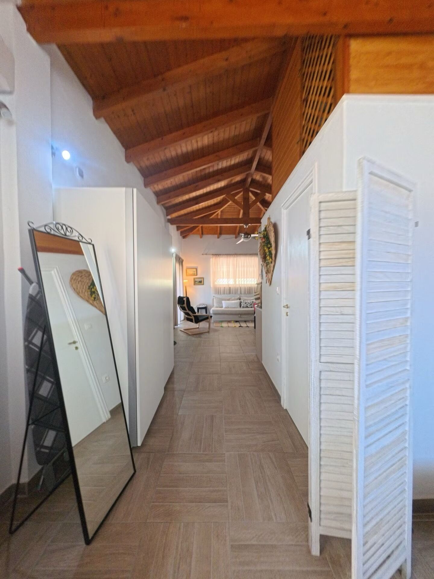 Un pasillo con techo de madera, paredes blancas y suelo de baldosas. Se ve un espejo apoyado contra la pared a la izquierda y una puerta blanca a la derecha.