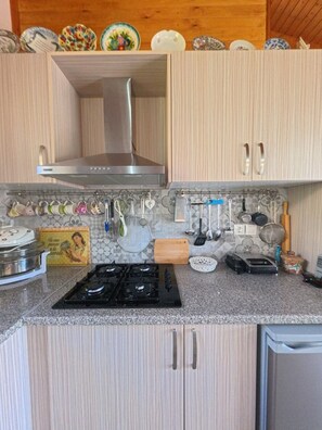 A kitchen with light wood cabinets, a stainless steel range hood, a gas stove, and various kitchen utensils hanging on the wall.