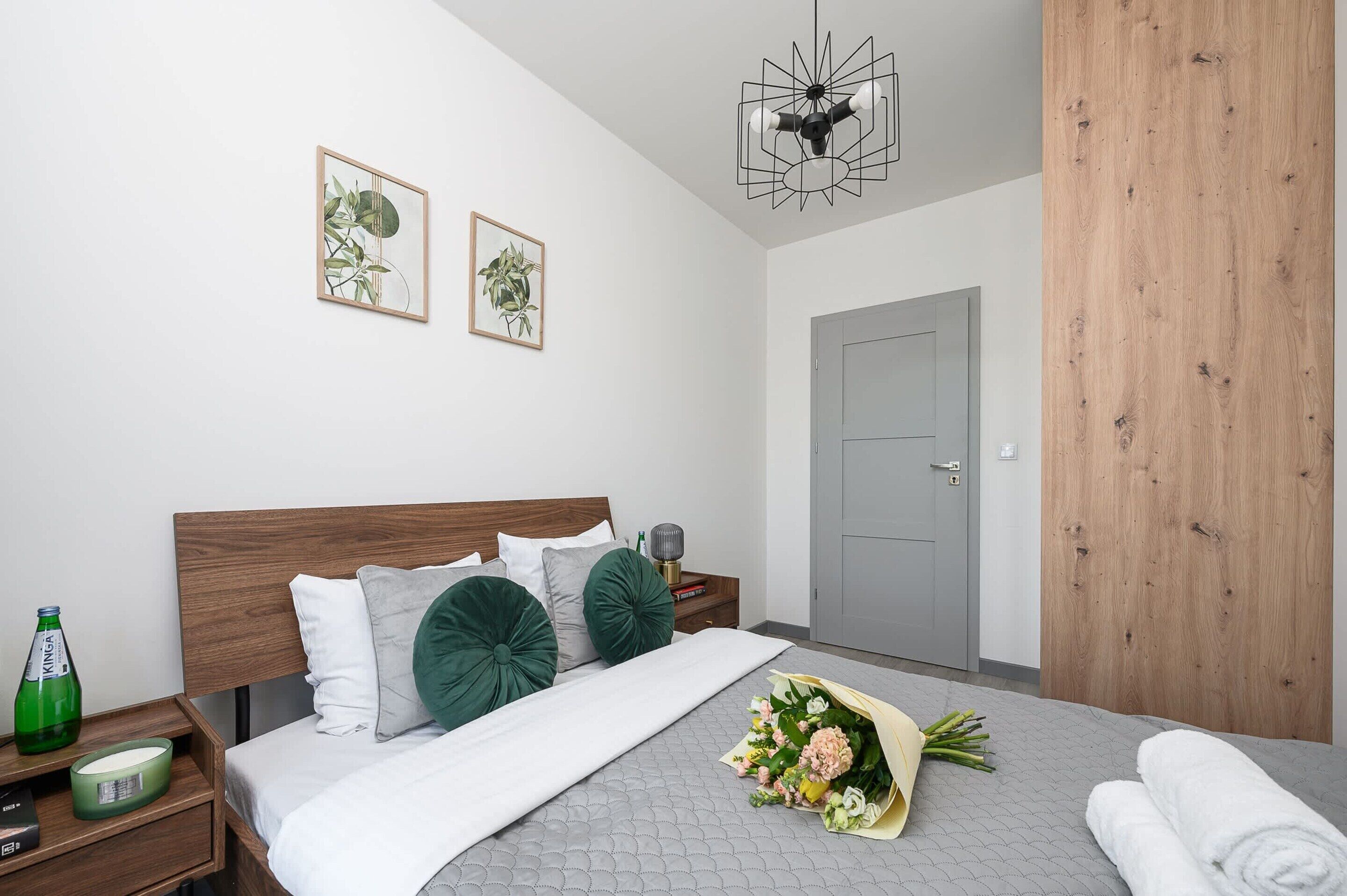 Een moderne slaapkamer met een houten hoofdbord, een grijs gewatteerd sprei, twee groene sierkussens, een boeket bloemen, een fles water en een groene kaars op een nachtkastje.