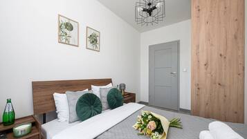 Un dormitorio moderno con cabecera de madera, una colcha gris acolchada, dos almohadones decorativos verdes, un ramo de flores, una botella de agua y una vela verde sobre una mesita de noche.