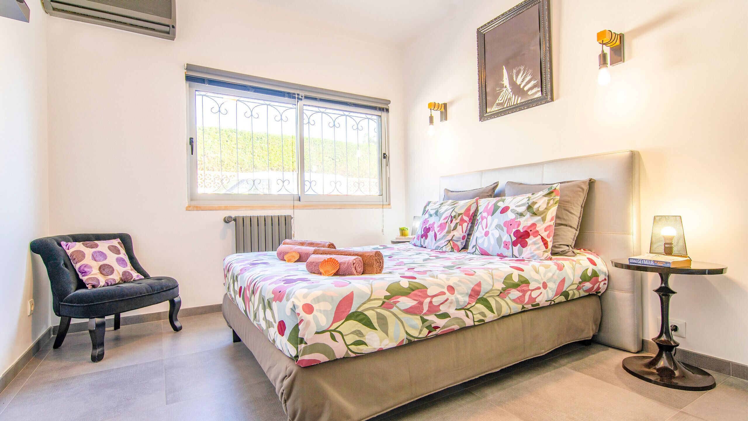 A bedroom with a bed, a chair, a radiator, a window with bars, a wall-mounted light, and a small table with a lamp.
