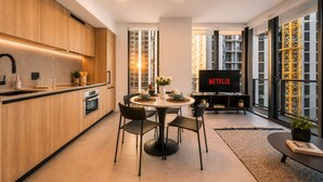 A modern kitchen with wooden cabinets, a dining area with a round table and chairs, a television displaying 'NETFLIX', and large windows with a view of a building. - Downtown Miami 1BR with City Views (Miami)