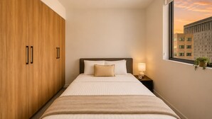 A hotel room with a bed, a nightstand, a window with a view of buildings, and wooden wardrobe doors. - Downtown Miami 1BR with City Views (Miami)
