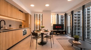 A modern kitchen with wooden cabinets, a dining area with a round table and chairs, a flat-screen TV, and large windows offering a view of a building.