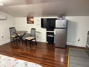 A compact living space with a dining table, two chairs, a refrigerator, a trash bin, and a television.