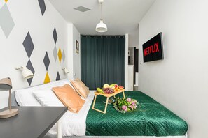 Ein Hotelzimmer mit einem Bett, einem kleinen Tisch mit Obst und Blumen, einem an der Wand montierten Fernseher und einem geometrischen Wandmuster.