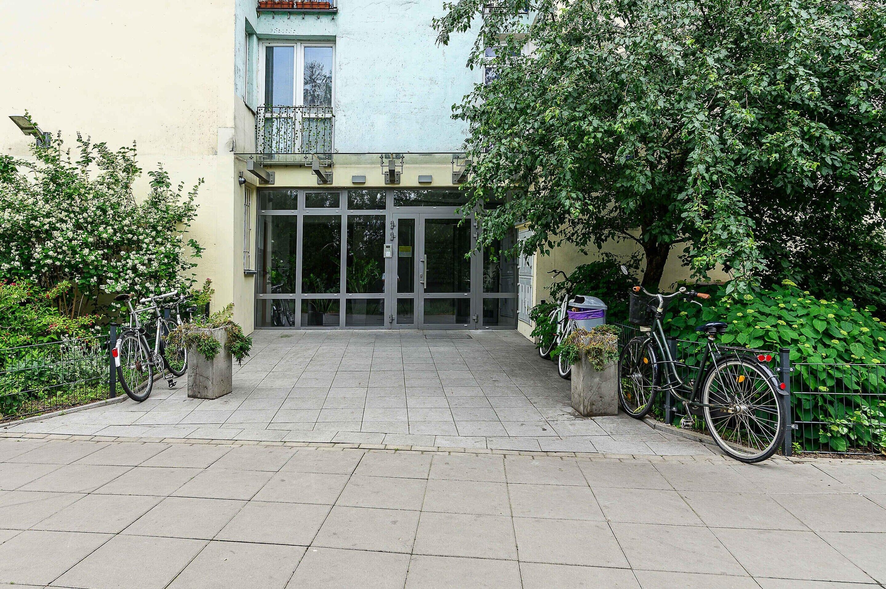 Ein Gebäudeeingang mit einem verschlossenen Tor, einem Fahrradstellplatz und einem kleinen Garten mit Blumentöpfen.
