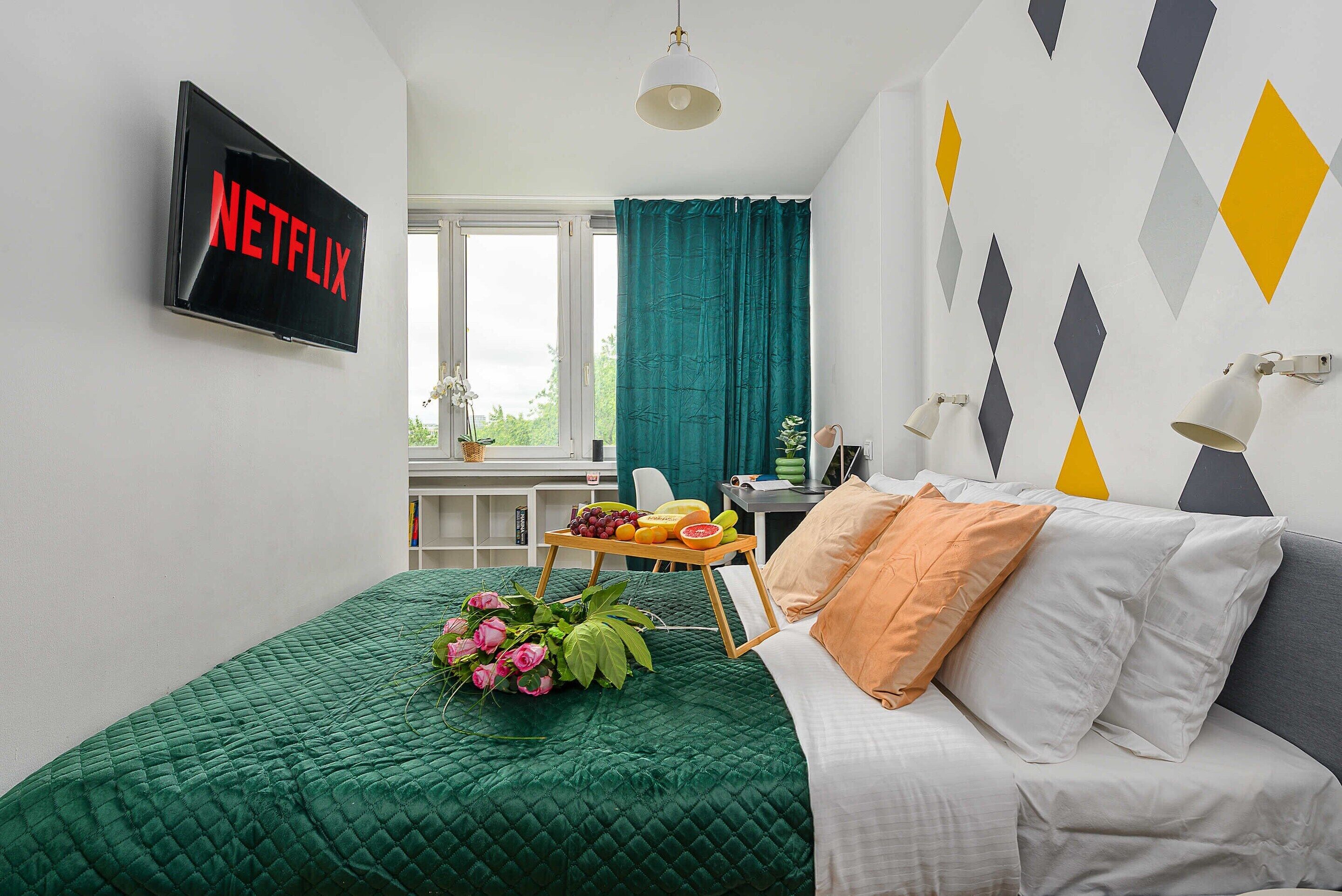 A modern bedroom with a green quilted bedspread, a white sofa with orange pillows, a table with fruit and flowers, a TV displaying 'NETFLIX', and geometric wall art.