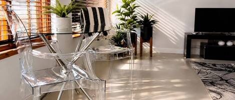 Una sala moderna con sillas transparentes, una toalla a rayas, plantas en macetas y un mueble para televisión.