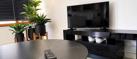 Una sala de estar moderna con un televisor de pantalla plana, una mesa de comedor redonda y dos plantas en macetas.