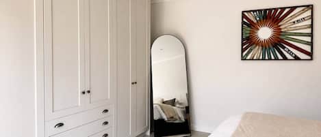 Quarto com guarda-roupa branco, espelho de corpo inteiro e uma obra de arte na parede.