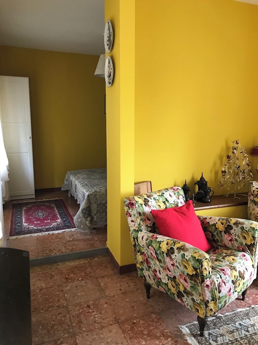 Een woonkamer met een fauteuil met bloemenprint, een rood kussen en een kamerbreed tapijt.