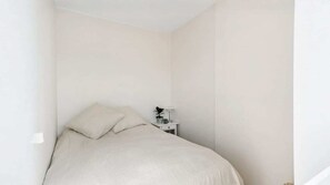 A small, single-bed room with a white bedspread and pillows, a nightstand with a lamp, and a potted plant. - Newly renovated and bright 2-bedroom apartment in the heart of Vesterbro, available for long-term rent (København)