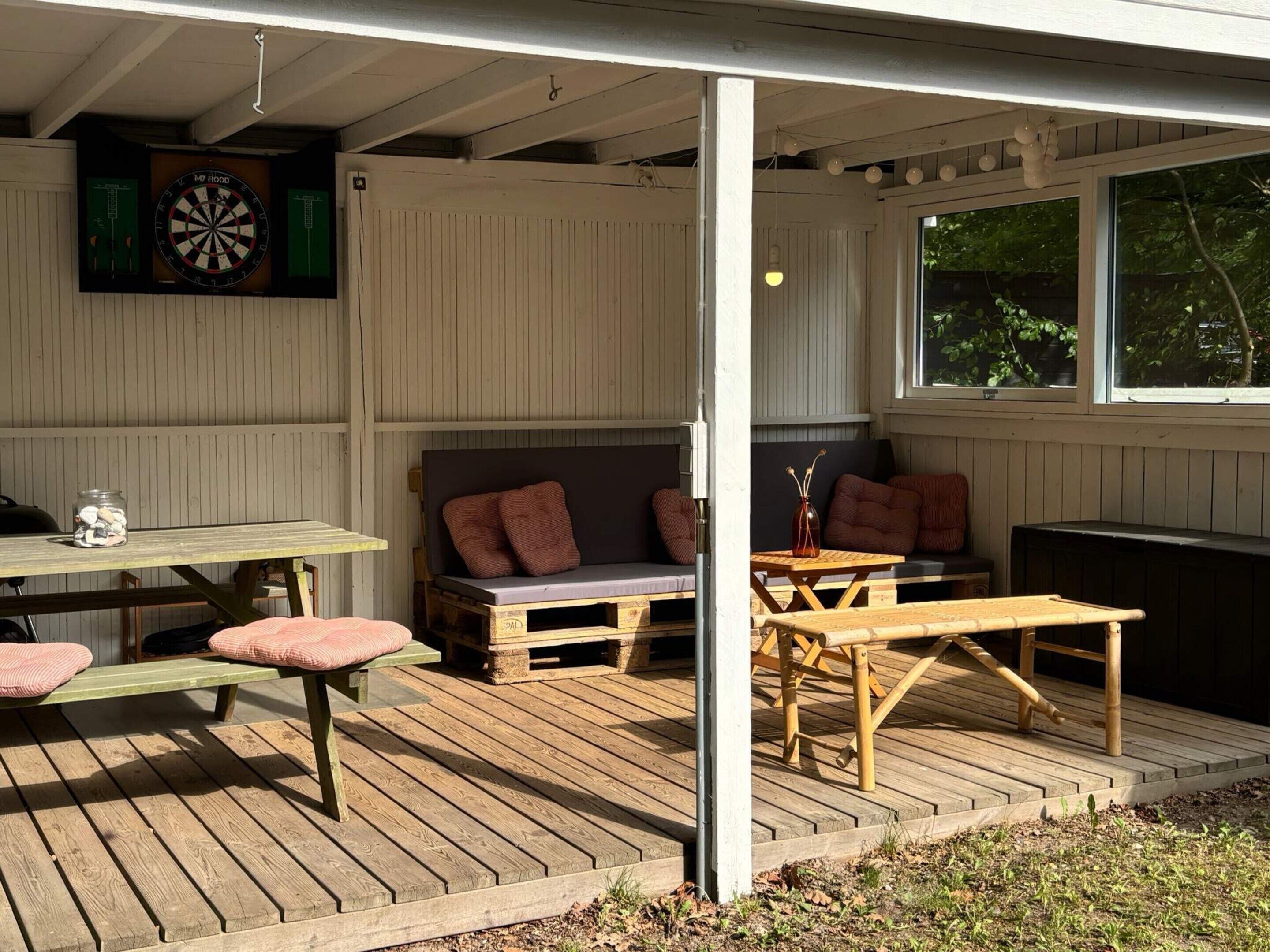 Une terrasse extérieure avec une table et des bancs, un jeu de fléchettes et une fenêtre donnant sur les arbres.