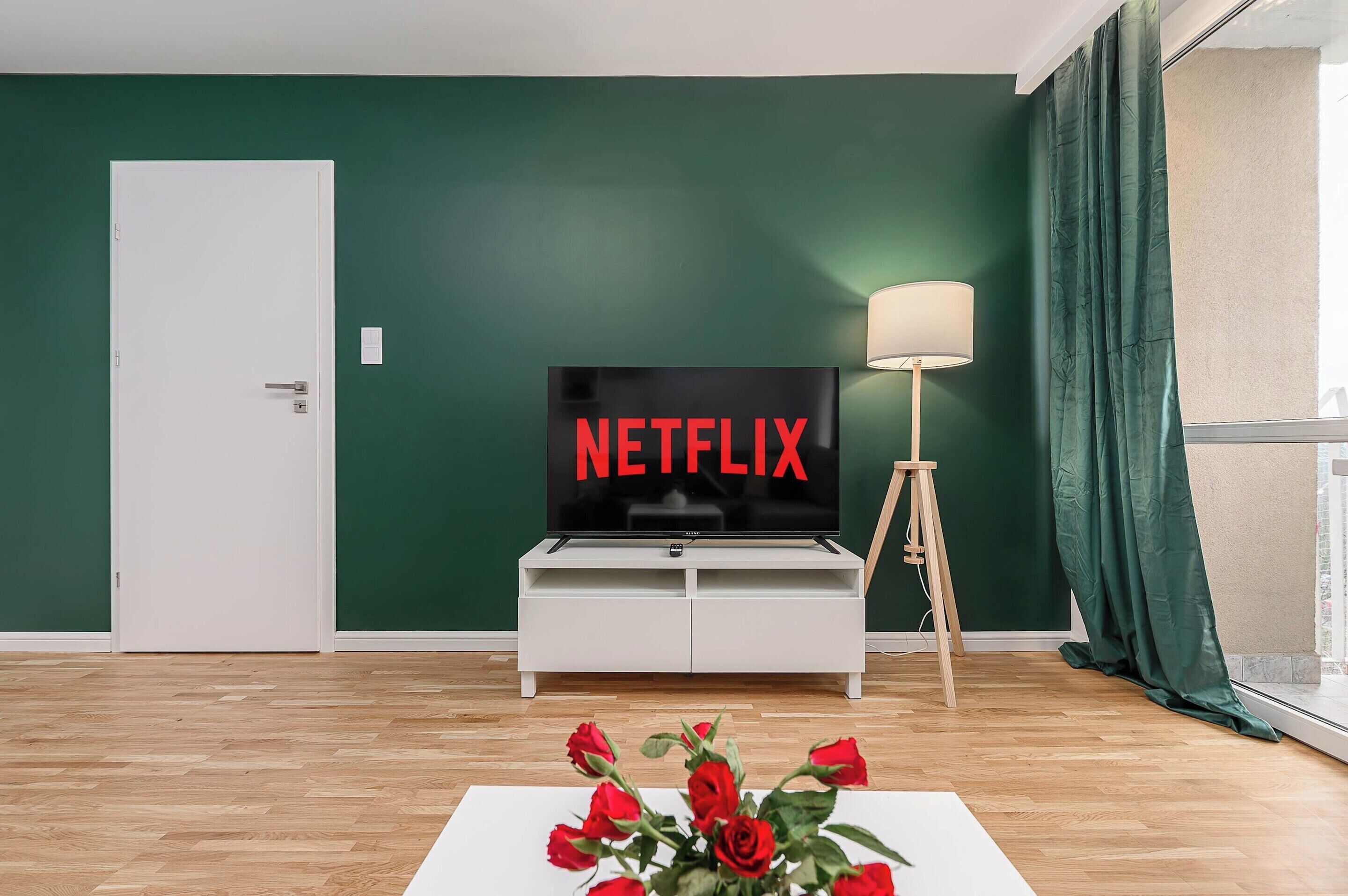 A living room with a television displaying 'NETFLIX' in red letters, a white cabinet, a floor lamp, and a vase with red roses.