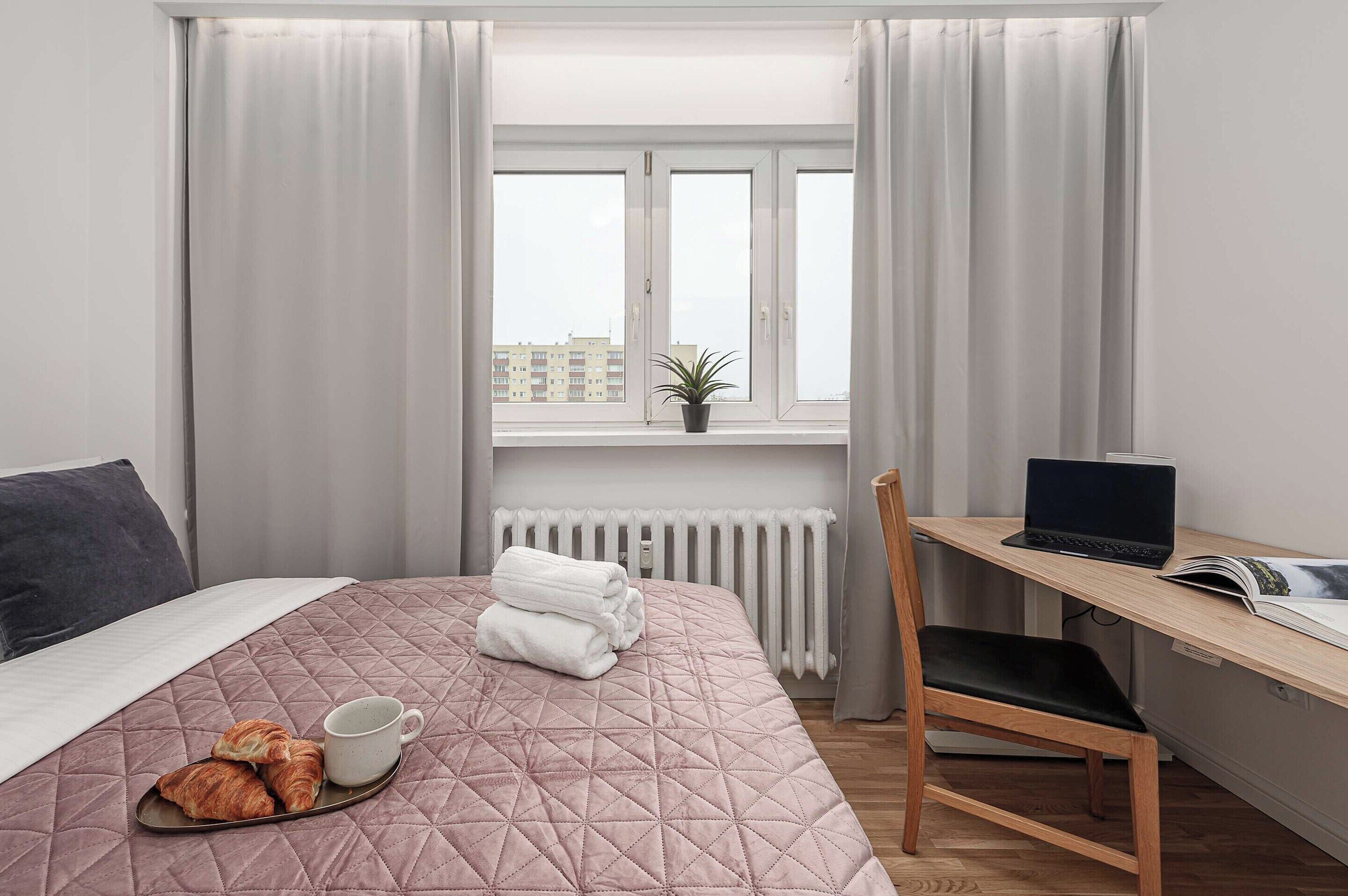 A hotel room with a bed, a desk with a laptop, a radiator, and a window with curtains.