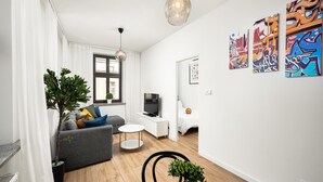 A modern living room with a grey sofa, a small round coffee table, and a TV on a white stand. There are plants on the floor and a large window with curtains. - Flat Near Piotrkowska Street (Łódź)