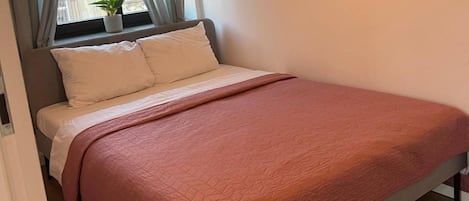 Una cama con colcha rosa, una ventana con cortinas y una planta en maceta sobre el alféizar.
