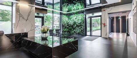 A modern lobby with a marble reception desk, seating area, and a large glass wall with a view of greenery.