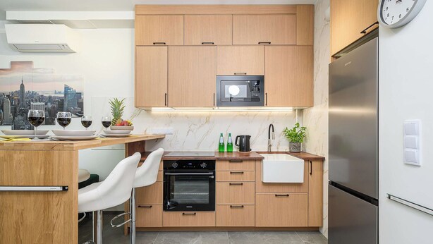 A modern kitchen with wooden cabinets, a built-in microwave, a stainless steel refrigerator, and a bar area with a white chair. - Lush Home With City Views (Warsaw)