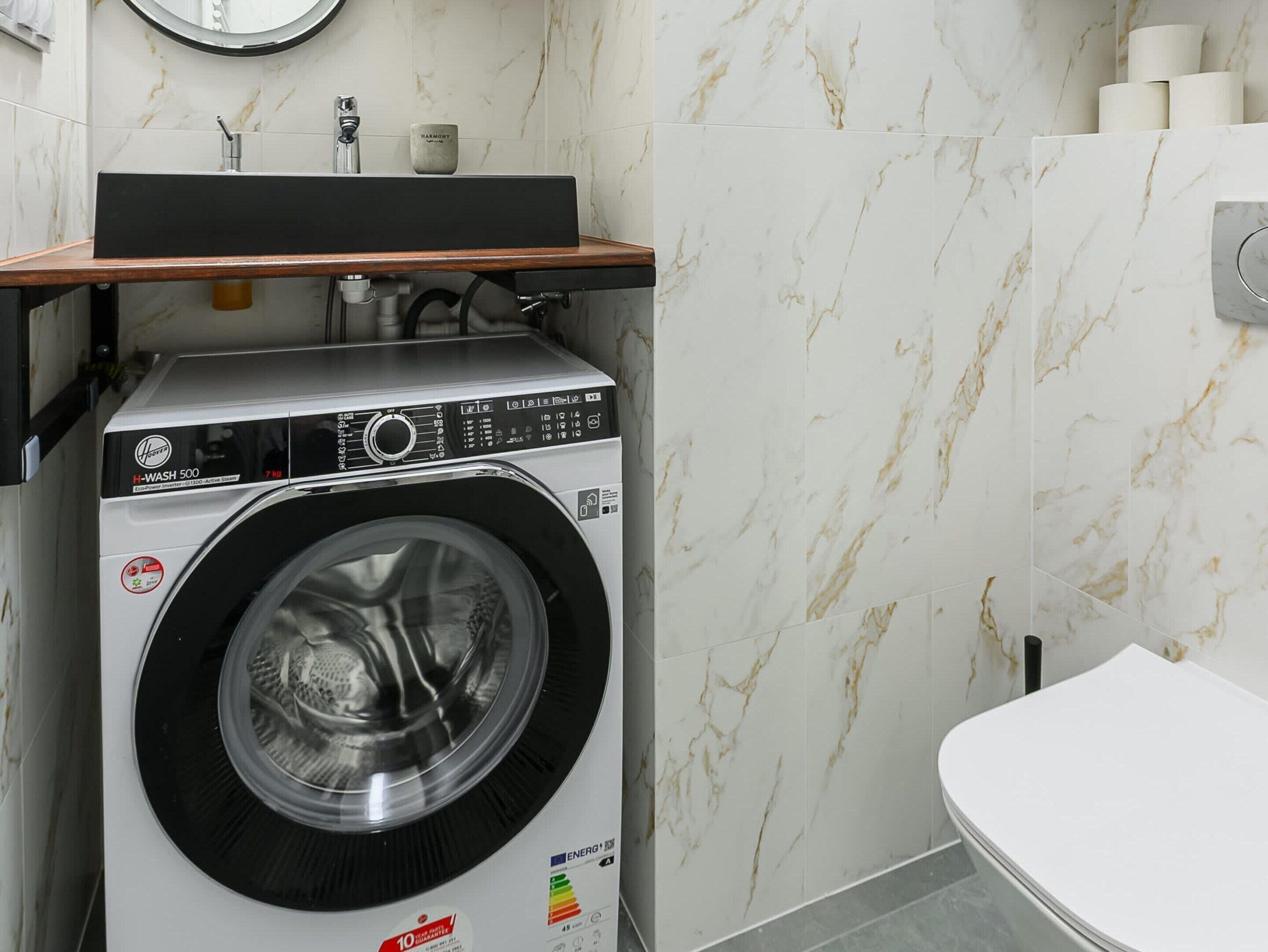A compact, modern laundry room with a front-loading washing machine, a small shelf above the sink, and a toilet to the right.