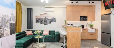 Un espacio de estar moderno con un sofá verde, una mesita redonda con un florero, una zona de cocina con armarios de madera, un refrigerador y un televisor de pared mostrando el logotipo de Netflix.