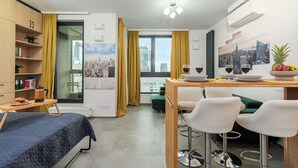 A modern hotel room with a bed, a dining area with a table and chairs, a bookshelf, and a cityscape view. - Lush Home With City Views (Warsaw)