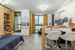 A modern hotel room with a bed, a dining area with a table and chairs, a bookshelf, and a cityscape view. - Lush Home With City Views (Warsaw)
