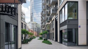 A modern urban street with high-rise buildings, a paved walkway, and small landscaped areas. - City Apartment With Balcony (Warsaw)