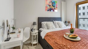 A modern hotel room with a large bed, a desk, a chair, a small table, a painting on the wall, and a window with a view of buildings. - City Apartment With Balcony (Warsaw)
