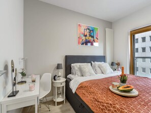 Ein modernes Hotelzimmer mit einem großen Bett, einem Schreibtisch, einem Sessel, einem kleinen Tisch, einem Bild an der Wand und einem Fenster mit Blick auf Gebäude.