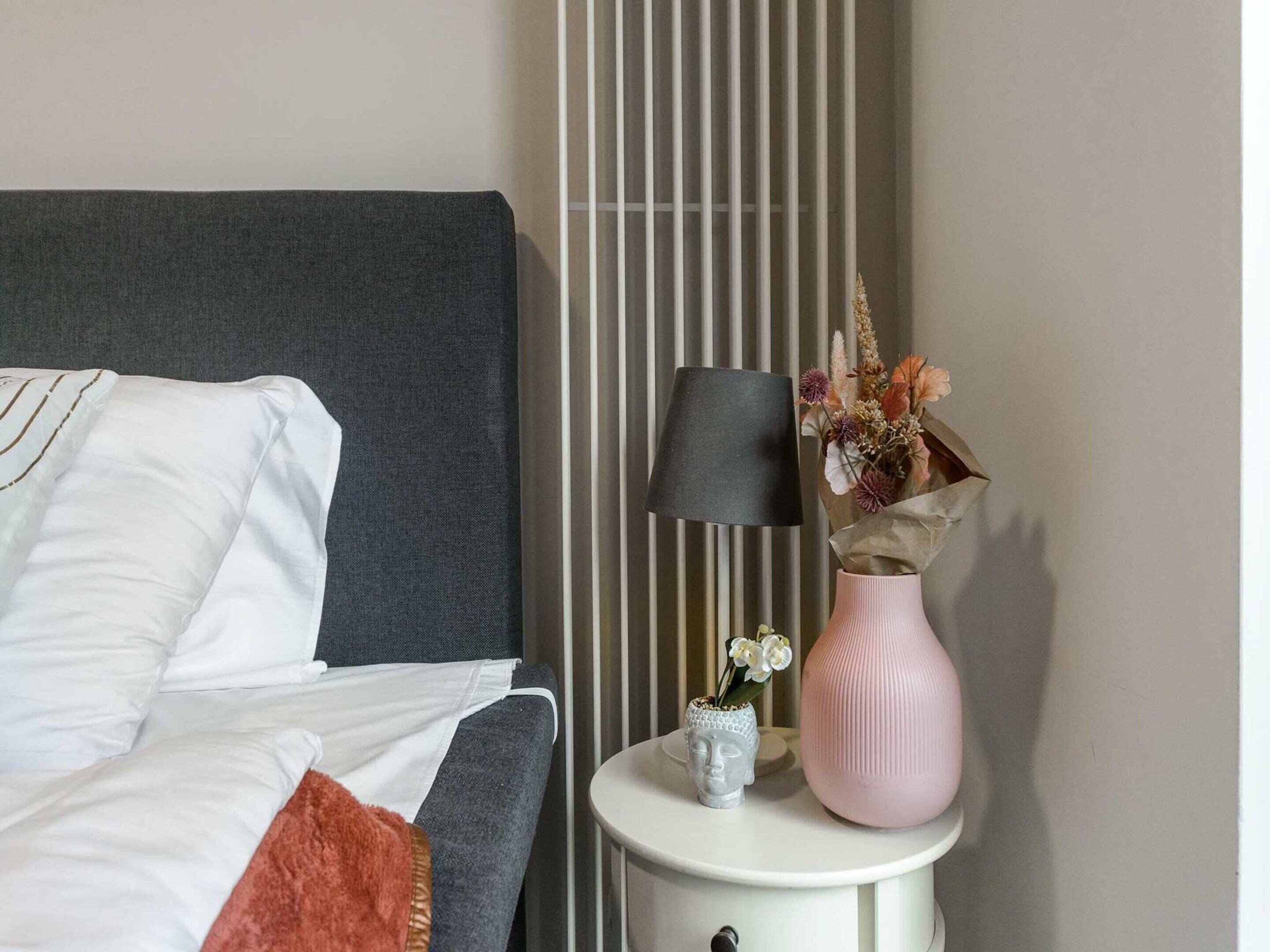 A neatly made bed with white bedding and a grey headboard, a bedside lamp, a vase with flowers, and a small Buddha statue on a bedside table.