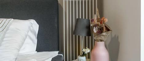 A neatly made bed with white bedding and a grey headboard, a bedside lamp, a vase with flowers, and a small Buddha statue on a bedside table.