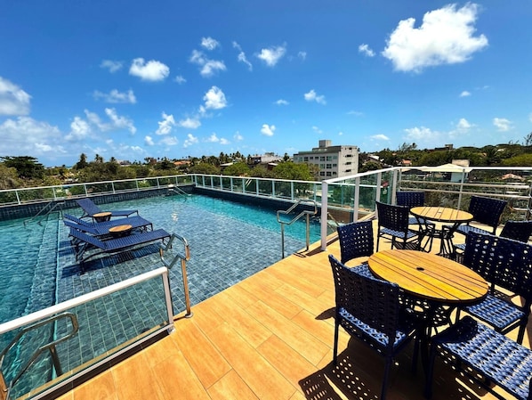 Uma área de piscina no terraço, com espreguiçadeiras e mesas, cercada por um guarda-vidro.