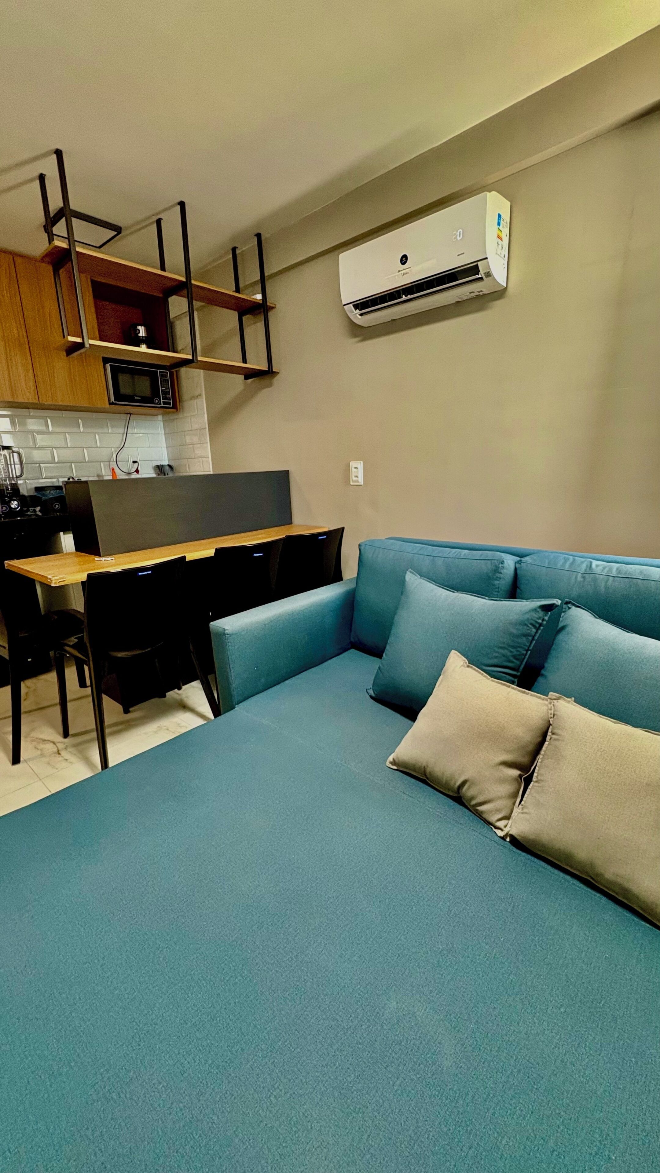 Um espaço de estar moderno com um sofá azul, uma mesa de jantar com cadeiras, cozinha com micro-ondas e um aparelho de ar condicionado.