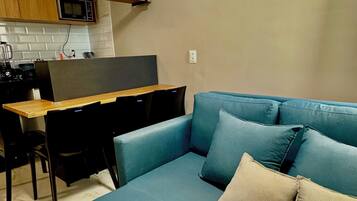Um espaço de estar moderno com um sofá azul, uma mesa de jantar com cadeiras, cozinha com micro-ondas e um aparelho de ar condicionado.