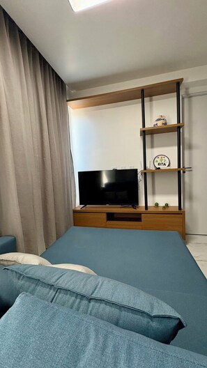 Quarto moderno com televisão de tela plana em um suporte de madeira, um sofá e uma prateleira fixada na parede com objetos decorativos.