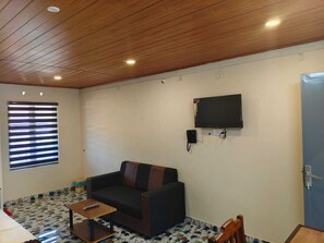 Una habitación con techo de madera, un sofá negro, una mesita, un televisor montado en la pared y una puerta.