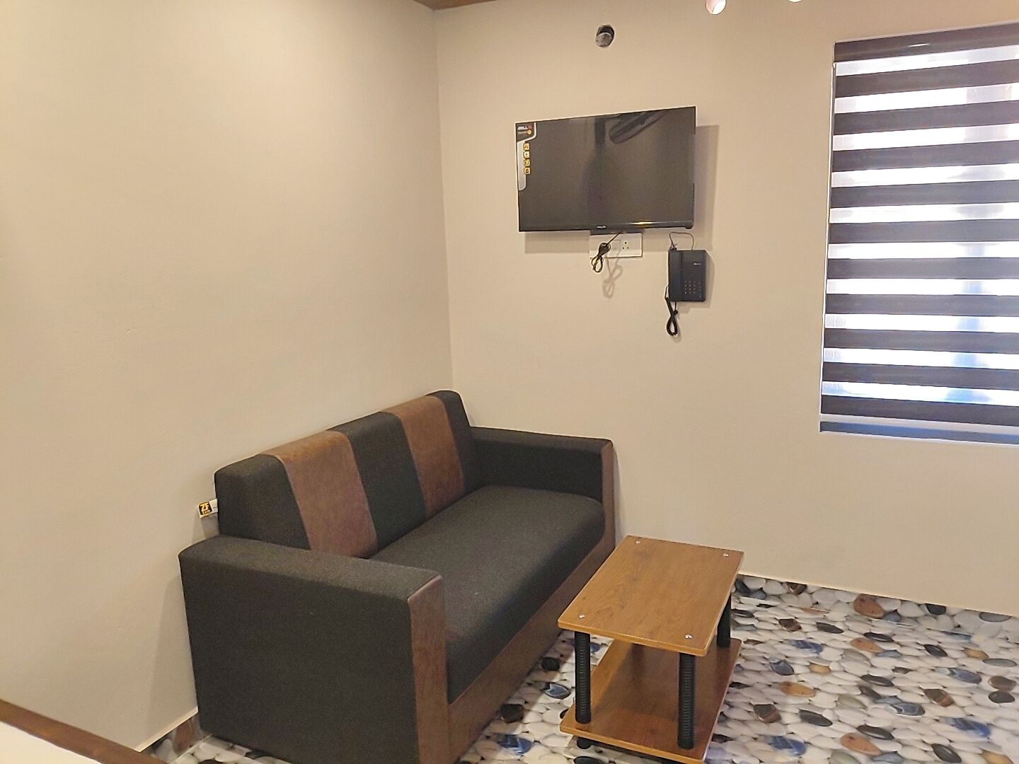 A living room with a sofa, a small wooden table, a flat-screen TV mounted on the wall, and a window with blinds.