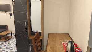 A compact room with a wooden dining table, a chair, a small kitchenette area, and a television mounted on the wall.
