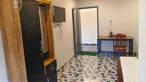 A hotel room with a wardrobe, a television mounted on the wall, a small table with a chair, and a pebble-patterned floor.