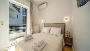 A hotel room with a bed, a bedside table, a lamp, and a view of a building outside. - Deluxe Apartment With Balcony and Views (Rethymno)